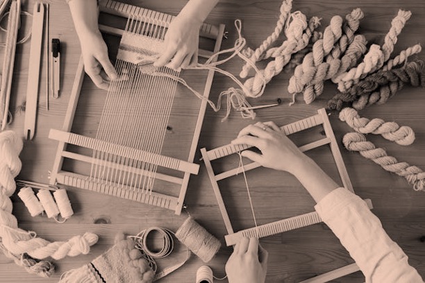 Looking down on two sets of hands hand weaving at looms with skeins of yarn on a table